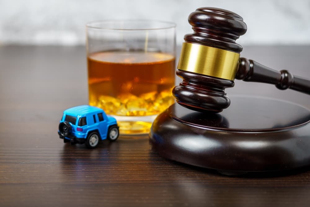 A judge's gavel rests on a courtroom table alongside a glass of whiskey and a miniature car, symbolizing the concept of "No drinking while driving."